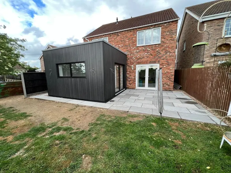 Modern single-storey rear extension clad in dark vertical boards, beside a red brick house and a new patio